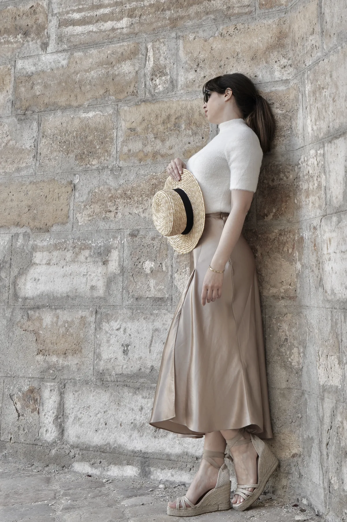 Model holding a straw hat against a stone wall, wearing wedge sandals.