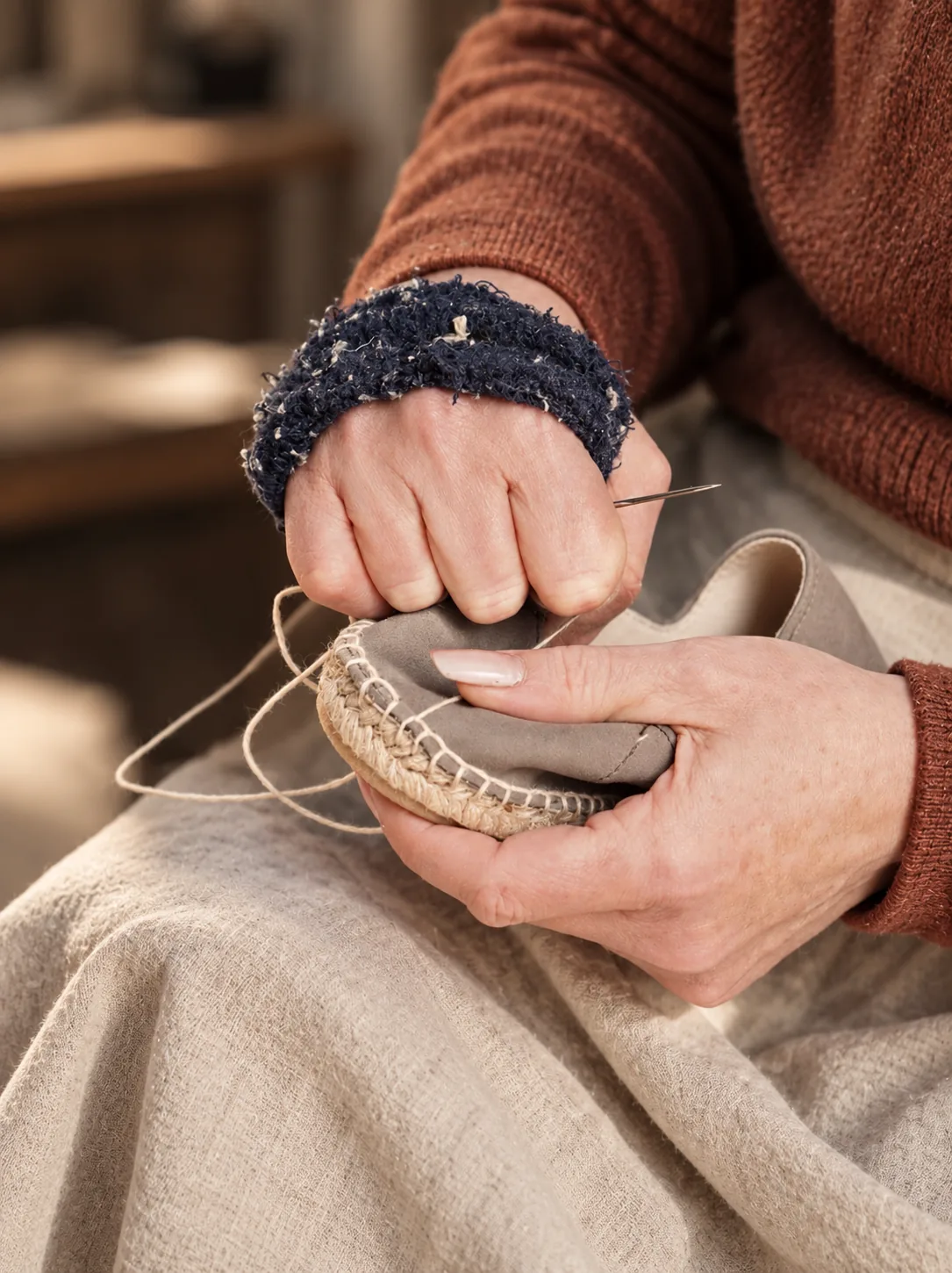 Hands stitching a grey suede upper to a jute sole with a needle and natural thread.