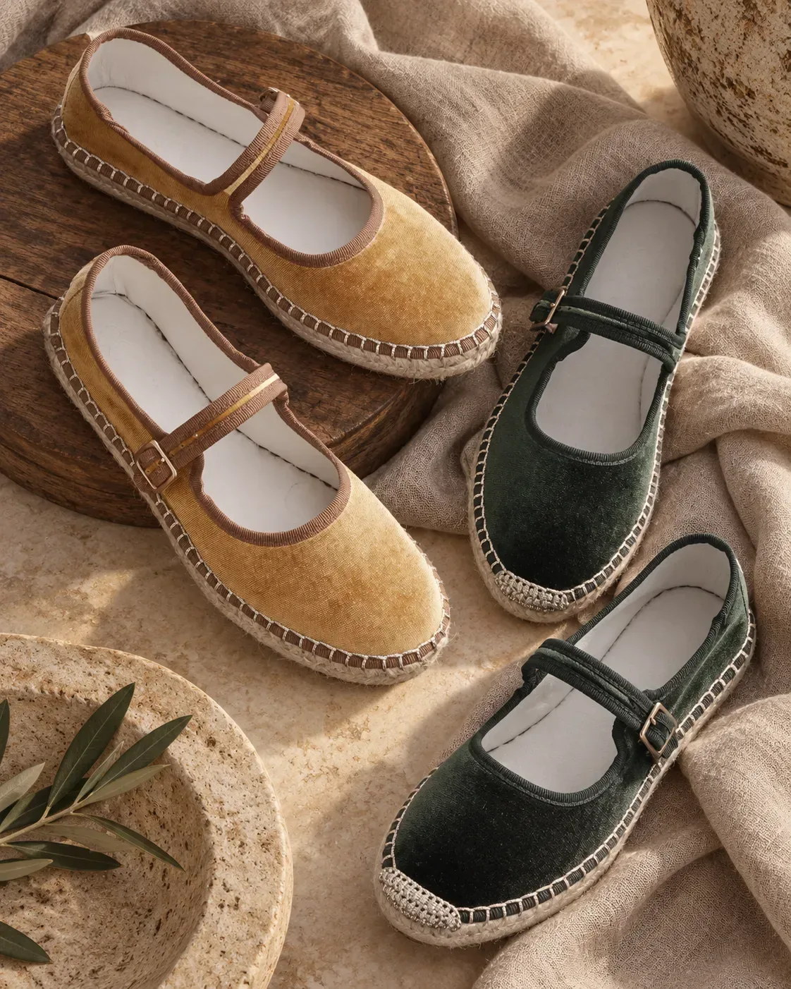 Two hand-stitched jute-sole Mary Janes in ochre and dark green velvet, resting on beige linen beside an olive branch.